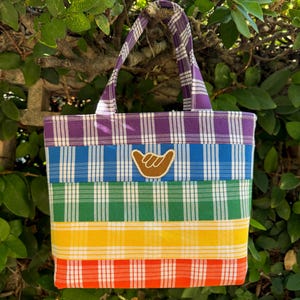 May include: A rainbow plaid tote bag with a brown shaka hand patch. The bag has a purple, blue, green, yellow, and red plaid pattern.