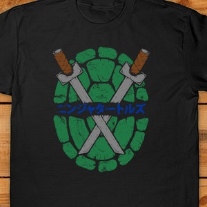 Peut inclure: T-shirt noir avec un motif de carapace de tortue verte, des &eacute;p&eacute;es crois&eacute;es et du texte japonais bleu. Le design a un aspect vieilli et vintage. Le t-shirt est en mati&egrave;re souple.