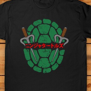 May include: Black t-shirt featuring a distressed green turtle shell design with two sai weapons and Japanese text. The design is centered on the shirt, creating a bold graphic.