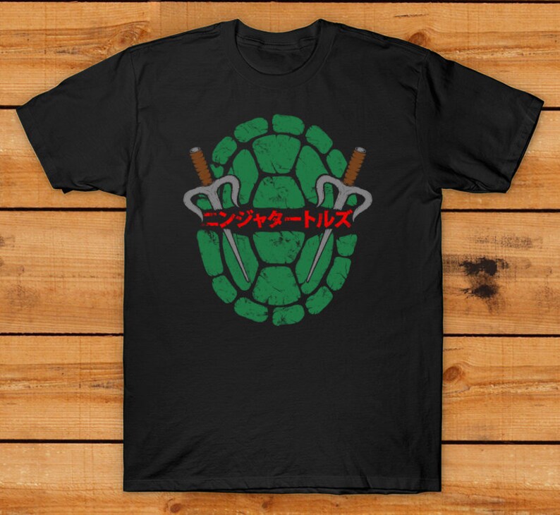May include: Black t-shirt featuring a distressed green turtle shell design with two sai weapons and Japanese text. The design is centered on the shirt, creating a bold graphic.
