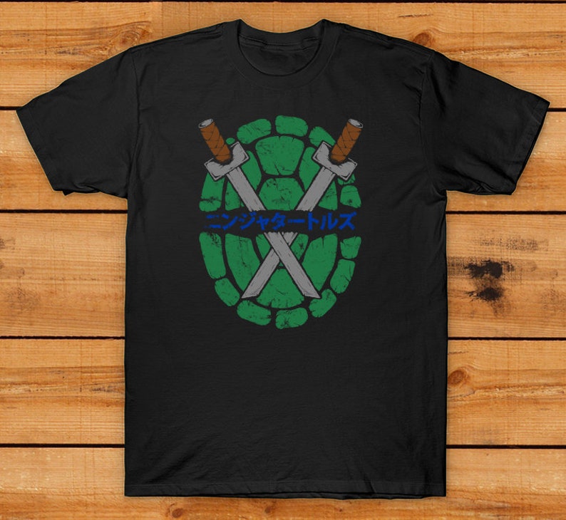 Peut inclure: T-shirt noir avec un motif de carapace de tortue verte, des &eacute;p&eacute;es crois&eacute;es et du texte japonais bleu. Le design a un aspect vieilli et vintage. Le t-shirt est en mati&egrave;re souple.