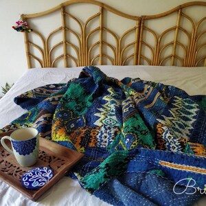 May include: A blue and green kantha quilt with a geometric pattern is draped over a white bed with a woven headboard. A blue and white ceramic mug sits on a wooden tray with a carved design.