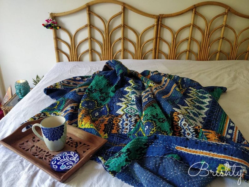 May include: A blue and green kantha quilt with a geometric pattern is draped over a white bed with a woven headboard. A blue and white ceramic mug sits on a wooden tray with a carved design.