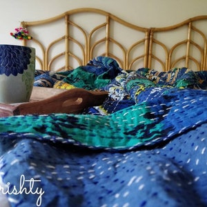 May include: A blue and green kantha quilt with white stitching is draped over a bed with a woven headboard. A white ceramic mug with a blue floral design sits on a wooden tray.