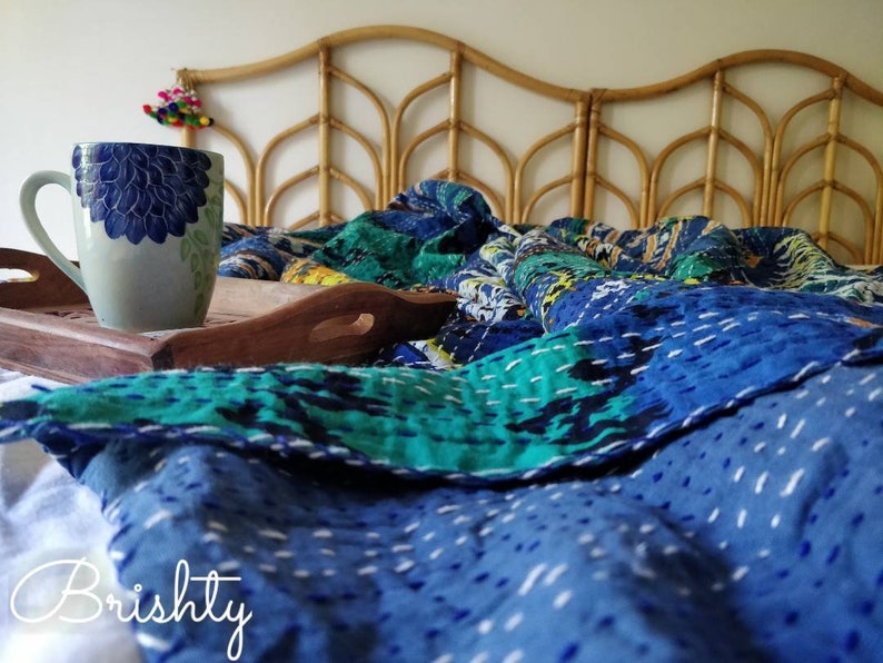 May include: A blue and green kantha quilt with white stitching is draped over a bed with a woven headboard. A white ceramic mug with a blue floral design sits on a wooden tray.