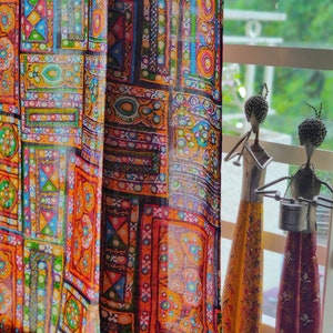Puede incluir: Una cortina colorida con un patrón geométrico en tonos de rojo, naranja, amarillo, verde, azul y morado. La cortina está hecha de una tela transparente y está colgada frente a una ventana. Dos figuras metálicas decorativas son visibles en el fondo.