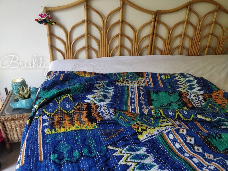 May include: A blue and green kantha quilt with a geometric pattern is draped over a bed with a light brown wicker headboard.