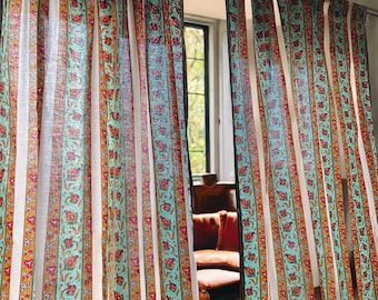 Puede incluir: Cortinas blancas con rayas de color turquesa, naranja y rojo y patrones florales. Las cortinas están colgadas en una ventana con vista a un césped verde.