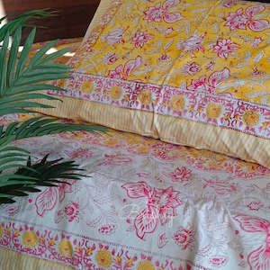 May include: A white cotton bed sheet with a pink and yellow floral print. The sheet has a yellow and white striped border. A yellow pillow with a pink floral print is on top of the sheet.