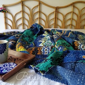 May include: A blue and green kantha quilt with intricate stitching, folded on a white bed with a woven headboard. A mug and coaster are on a wooden tray in the foreground.