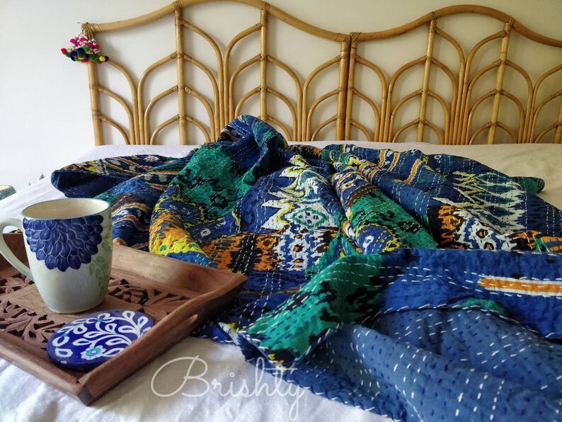 May include: A blue and green kantha quilt with intricate stitching, folded on a white bed with a woven headboard. A mug and coaster are on a wooden tray in the foreground.