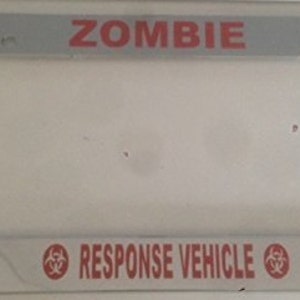 May include: A chrome license plate frame with the words "ZOMBIE RESPONSE VEHICLE" in red lettering. The frame has a biohazard symbol on each side.