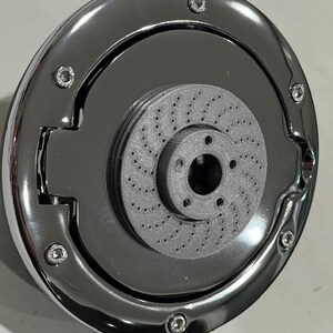 May include: A shiny chrome-plated automotive part with a circular design. The part features a central gray disc with numerous small holes, surrounded by a polished metal ring secured with small bolts. The overall design is symmetrical and industrial.