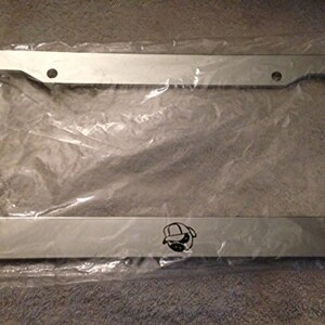 Puede incluir: Marco de matrícula de metal plateado con un gráfico en blanco y negro de una persona con sombrero y gafas de sol.