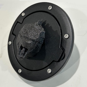 Puede incluir: Tapa de gasolina negra con una cabeza de Godzilla impresa en 3D en el centro. La tapa tiene un acabado texturizado y tornillos plateados alrededor del borde.