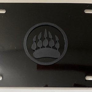 May include: Black license plate with a bear paw print design. The paw print is in a circular frame. The license plate has four mounting holes and a glossy finish.