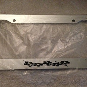 Puede incluir: Un marco de matrícula de coche plateado con un diseño floral negro. El marco tiene dos agujeros en la parte superior y dos agujeros en la parte inferior para el montaje.