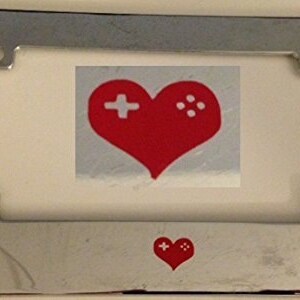 May include: Chrome licence plate frame with a red heart and a white plus sign and four white dots inside the heart. The heart is a symbol for video game lovers.