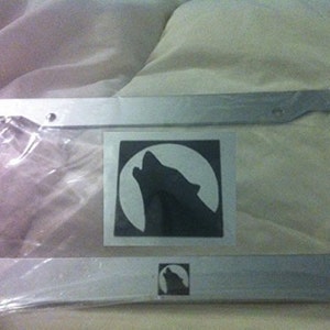 Puede incluir: Un marco de matrícula de metal plateado con un lobo negro aullando a la luna. El marco tiene una pequeña silueta de lobo en la esquina inferior derecha.