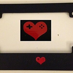 May include: Black licence plate frame with a red heart and a video game controller design. The frame has two holes on each side for mounting.