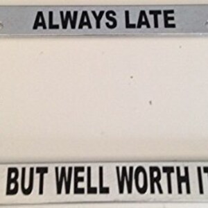 May include: A silver metal license plate frame with black text that reads "ALWAYS LATE BUT WELL WORTH IT".