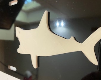 May include: A white plastic shark silhouette attached to a black surface. The shark is facing the left side of the image with its mouth open.