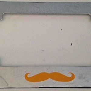 May include: A chrome licence plate frame with a bright orange moustache design.