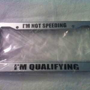 May include: A silver license plate frame with the text "I'm not speeding, I'm qualifying".