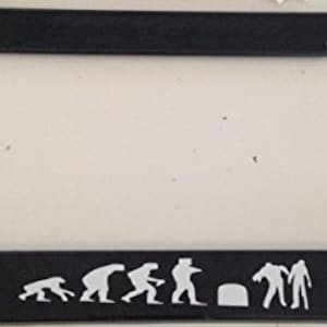 May include: Black license plate frame with a white silhouette of the evolution of man, ending with a zombie. The text reads "_"
