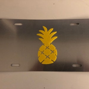 Puede incluir: Una placa de matrícula de metal plateado con un diseño de piña amarilla.