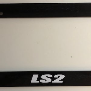 May include: Black plastic license plate frame with white text that reads "LS2".