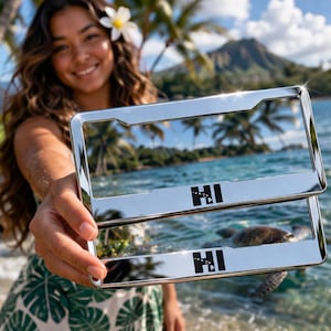 Puede incluir: Dos marcos cromados brillantes para matrículas con las letras "HI" en negro. Los marcos se sostienen frente a una escena de playa tropical con palmeras, océano y una montaña al fondo.
