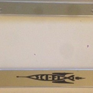 Op de afbeelding: Een glanzend, zilveren kentekenplaatraam. Het frame heeft een rechthoekige vorm met afgeronde hoeken en montagegaten. De onderkant van het frame heeft een zwarte afbeelding van een raket met de tekst "SCI-FI".