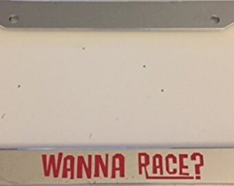 Op de afbeelding: Een zilveren kentekenplaathouder met een rode en witte sticker waarop "WANNA RACE?" staat.