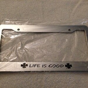 Puede incluir: Marco de matrícula de coche plateado con el texto "LIFE IS GOOD" y un trébol a cada lado.