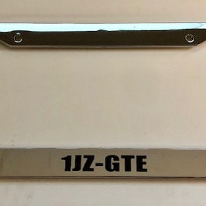 May include: A chrome licence plate frame with the text "1JZ-GTE" printed in black.