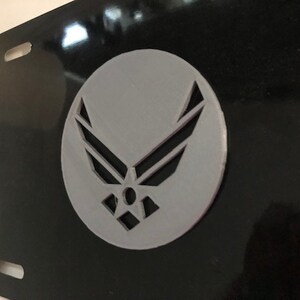 May include: A black license plate featuring a gray emblem of the United States Air Force. The emblem is a circular design with stylized wings and a star in the center. The plate has two mounting holes.
