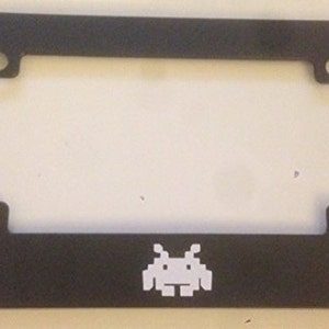 May include: Black metal license plate frame with a white pixelated alien graphic.