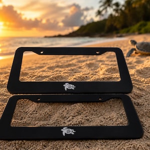May include: Two rectangular black license plate frames featuring a white sea turtle graphic centered at the bottom edge. Each frame has two mounting holes at the top and a smooth matte finish, designed to hold a standard vehicle license plate.