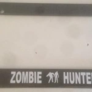 May include: A black plastic license plate frame with the text "ZOMBIE HUNTER" in white letters and a silhouette of two people in the middle.