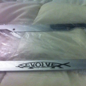 May include: Silver license plate frame with the word "EVOLVE" and a fish graphic in black. The frame is rectangular and appears to be made of metal. The frame is in a clear plastic bag.