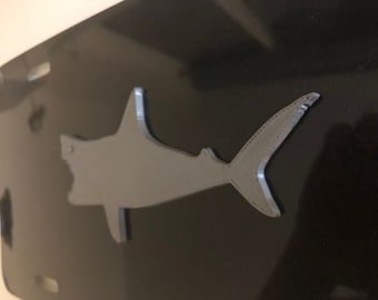 May include: A black metal license plate with a gray metal shark silhouette.