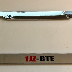 May include: Chrome license plate frame with a white background and the text "1JZ-GTE" in red letters.