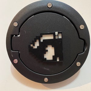 Può includere: Un tappo del serbatoio rotondo nero con una vite argentata attorno al bordo e un'immagine pixelata bianca di un personaggio di videogioco al centro.