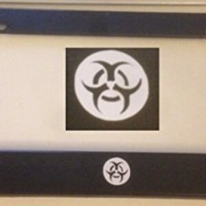 May include: Black license plate frame with a white biohazard symbol in the center and at the bottom. The frame is rectangular with rounded corners and holes for mounting. The biohazard symbol is a white graphic on a black background.