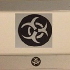 May include: A silver license plate frame with a black biohazard symbol in the center. The symbol is a stylized three-pronged design.