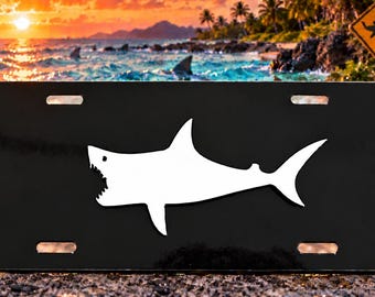 May include: A rectangular black license plate featuring a centered white silhouette of a shark with its mouth open. The plate has four mounting slots, two along the top edge and two along the bottom edge, and a smooth, glossy finish throughout.