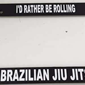 May include: Black license plate frame with white text that reads "I'd rather be rolling Brazilian Jiu Jitsu".