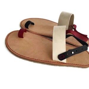 Puede incluir: Dos pares de sandalias de cuero con correas ajustables. Las sandalias tienen un bucle para el dedo del pie y una correa trasera. Las correas son de diferentes colores, incluyendo negro, rojo y beige.