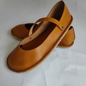 Pode incluir: Um par de sapatos baixos de couro castanho com um detalhe de costura vermelha em volta da sola. Os sapatos t&ecirc;m um design simples com uma &uacute;nica tira na parte superior.